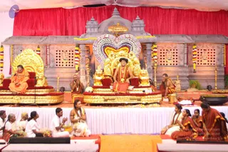A photograph of a satsang on a raised stage draped in red fabric, with a grey stone temple-style backdrop. A seated man in orange-red robes, a dark cap and a gold necklace sits on an ornate golden throne with lion motifs and a large multicoloured arch behind him, smiling as he addresses the gathering. A second seated figure in orange robes sits on a smaller golden throne to the left, and two monks in orange sit nearby. A gold kalasha stands at the right edge beside floral garlands on the backdrop. In the foreground, devotees in white and red-bordered saris and white dhotis sit on white cloth, some with hands folded in namaskar, and a man in white holds a musical instrument. Marigold garlands and lamps decorate the stage.