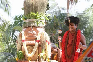 Outdoor ritual photograph in a palm-fringed garden. A tall carved stone or terracotta kalasha-like pot, topped with a bundle of dried grass and green leaves and wrapped in a rope, is decorated with white jasmine garlands and red flower garlands. A standing man in a dark turban-like crown with a gold ornament and a bright red rose garland, wearing an orange-red robe and a brown rudraksha mala, smiles while holding a silver incense or lamp vessel and a small silver bell. Beside him, a seated or bent man in an orange-red cloth and a rudraksha mala looks down near the base. a person is the standing man in the orange-red robe with the ornate crown and garlands, described by his crown, saffron-toned clothing and rudraksha malas; the bent man is a devotee or priest.