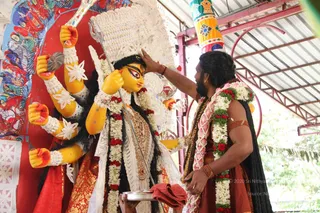 A man in a rust-orange dhoti and shawl, wearing a pink-and-white flower garland, long dark hair and a beard, presses his right hand to the forehead of a yellow-faced deity murti, a goddess with multiple arms holding a trident, mace and other implements, adorned with a white and gold crown, white garlands with red flowers and a gold-embroidered red sari. A brass plate with a red cloth sits at the base. Behind them a painted red and multicolour backdrop and a corrugated metal roof frame the scene.