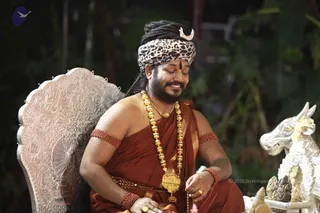 A seated man in a rust-brown saffron-toned robe with a gold-coloured shoulder border sits in an ornate silver-carved throne with floral scrollwork. He wears a leopard-print turban with a curved white horn-like ornament at the top, a red tilak on his forehead, rudraksha-style red beaded armlets, and a long gold bead necklace with a large gold pendant. His eyes are closed in a serene smile as he holds a prayer bead mala. To the right, a silver carved bull or nandi figure with a garland stands near a small arrangement of silver ritual objects. A dark leafy background frames the scene. Visible copyright credit text appears in the lower corner.
