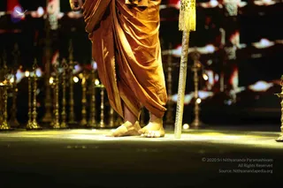 Close-up photograph of a standing figure's bare feet and lower body on a dark stage lit by a warm spotlight. The figure wears a rust-orange draped dhoti with a gold border and holds a tall silver staff with a gold tassel at the top. Behind are rows of gold oil lamps on stands and a blurred red and white backdrop. Only the legs and feet are visible; no face or full body appears in the frame.