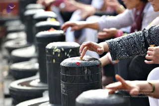 A row of black stone Shivalingams stands in the foreground, their tops wet and marked with yellow turmeric and red kumkum. Several devotees sit behind them, reaching out to touch and anoint the lingams during a Rudra Abhishekam. In the foreground, a woman in a black and white patterned long-sleeved top extends her hand to a lingam, and another person's hand appears at the right edge. Others in white and grey clothing sit in a line beyond. No a person candidate is visible; the people are shown only from the arms and torsos down or in profile.