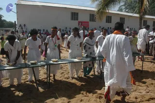 Photograph of a sandy outdoor festival setting with a row of plain metal tables holding small brass bowls. Young people in white clothes and orange headbands stand along the tables, some with white shawls. A man in a white kurta with an orange cloth over his shoulder stands at the right, facing the table. Behind them a long white building with a corrugated roof and palm trees, and a crowd of onlookers in white and colourful clothes gathers. A red banner with a portrait is on the wall. No identifiable a person appearance is visible in this frame.