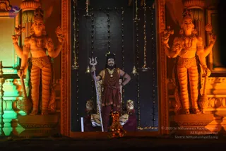 A photograph of a central figure standing in a dark recessed alcove framed by two large ornate golden-orange statues of multi-armed deities with crowns and jewelry. The central figure wears a maroon-brown robe with gold trim, layered rudraksha malas and a gold ceremonial necklace, holds a silver trident in one hand, and stands beside a gold-tiered seat draped with a tiger-striped cloth. Strings of small lights and hanging bells decorate the dark backdrop, and a golden oil lamp sits at the base. Green and orange stage lighting colours the surrounding structure.