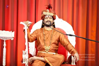 A photograph of a man seated cross-legged on an ornate white and red throne against a red draped backdrop. He wears a red-and-gold turban with a golden ornament, orange-brown silk kurta, multiple rudraksha malas and a large gold pendant necklace, and gold earrings. He holds a tall white ceremonial staff with a lion-shaped base in one hand while gesturing, and speaks into a microphone on a stand. A white carved lectern stands at left.