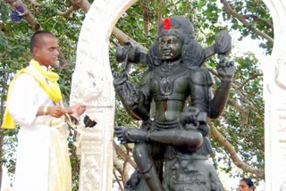 A photograph of a dark stone deity murti, a four-armed Shiva-form figure seated and adorned with a red flower above the crown, being bathed with water running down its chest and arms. A shaven-headed priest in white dhoti and a yellow shawl stands at left, holding a small brass vessel and a cloth, apparently performing abhishekam. The statue is framed by a carved white stone archway with green leafy trees behind. A woman's head is partially visible at lower right.
