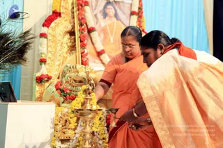Two women in orange and saffron saris perform an aarti or lamp offering before a golden throne decorated with red and yellow flower garlands and a peacock-feather fan. On the left, a woman in orange with glasses holds a brass lamp steady; on the right, a woman in an orange-and-gold bordered sari bends forward, lighting or tending the lamp. A small brass deity figure sits among marigold garlands on the altar, and a framed portrait of a man in orange robes with a white bead necklace is garlanded behind the throne, its image labelled as a portrait. Blue drapes frame the stage.