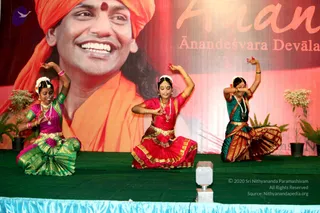 Three young female dancers perform a classical Indian dance on a green-covered stage. They wear saris and costume jewellery: the left dancer in magenta and green with a green-and-gold border, the centre dancer in red and pink with gold trim, and the right dancer in deep teal with gold border. Their arms are raised in mudra-like poses, with flowers in their hair and necklaces. Behind them a large backdrop shows a smiling man in orange robes and an orange head wrap, with the large printed phrase Ananda and the caption Ānandeśvara Devālaya in orange lettering. Potted plants flank the stage. The man on the backdrop is a printed portrait, not a physical person present.