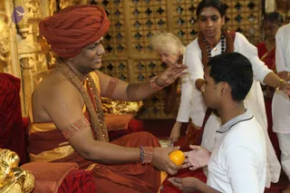 A seated man in an orange-red turban, rust and gold robes, a gold beaded necklace and beaded bracelets sits on a red cushioned throne before an ornate gilded wooden lattice backdrop. He extends his hand to bless a kneeling young man in a white shirt, who holds out his palms to receive an orange fruit. Behind them a woman in white with a sandalwood tilak and a rudraksha mala stands watching, and an older woman in white and others in red and white stand nearby. The scene suggests a satsang initiation moment.