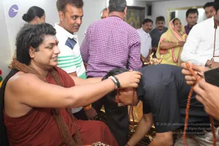 A photograph inside an indoor room where a seated man in a rust-red robe with long rudraksha mala beads and a shaved-looking dark wavy hairstyle places his hand on the head of a man bent forward in a dark shirt. The seated figure has a red bindi and a beaded bracelet and wears a wide rudraksha necklace draped over his shoulders, appearing to give a blessing. Around them stand several onlookers: a man in a striped white polo, a man in a purple checked shirt, a woman in a yellow sari with pink border, and men in white and grey shirts, one holding a phone. A brass pot and a golden object sit near the floor, and a framed picture hangs on the wall behind.