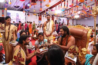 A festive hall decorated with hanging multicoloured lanterns and a large screen showing a bearded man in saffron. In the foreground a bearded man with long hair, wearing an orange-saffron garment, rudraksha mala and a brown-beaded necklace, sits on an ornate gold-trimmed brown leather throne, holding a silver-framed devotional card with a sacred image. Around him devotees offer similar framed cards; a woman in a gold saree and a woman in green stand nearby, a man in white dhoti with a gold border and mala stands behind the throne, and a gold trident rests on the right. Many seated and standing devotees fill the red-carpeted floor, some in saris and salwar kameez.