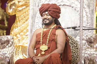A smiling man in saffron-orange clothing and a matching saffron turban sits on a silver-and-gold throne with a leopard-print cushion behind him. He wears a large gold ceremonial necklace with a gold pendant, a red tilak on his forehead, and long dark hair swept back. His hands are clasped in his lap, and a microphone stand is positioned before him beside silver lion statues. Gold ornate decorations fill the background.