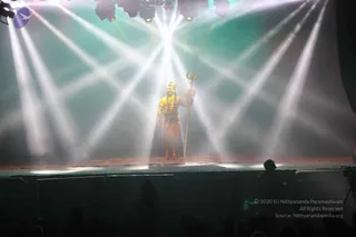 A figure in an orange-gold costume with a trident stands centrally on a dimly lit stage beneath sweeping white spotlight beams. The figure, shown in profile with long dark hair and bare upper body with adornments, holds a golden trident upright. The stage floor is red and gold. Audience members appear as dark silhouettes in the foreground, and a camera operator sits at the right edge. The scene resembles a theatrical stage performance, possibly a costumed Shiva figure.