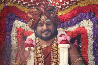 A photograph of a smiling man seated before a dense backdrop of red, pink, purple and white flower garlands. He wears a rust-brown turban with gold ornaments and green beads, a gold forehead piece, a thick gold necklace of beaded pendants, a garland of white jasmine and pink and red blossoms, and a rust-coloured garment. He raises one hand in a blessing gesture, with gold rings and gold bangles visible.