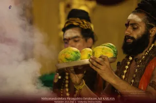 Two men in ritual dress perform a homa in a temple setting, with white smoke rising at the left edge of the frame. The man in front, bearded with long hair, white forehead markings and a red tilak, wears a brown shawl, orange cloth and layered gold beaded necklaces, and holds a silver tray of ripe yellow-green fruits toward the fire. Behind him, a second man with a striped yellow-and-black headwrap and white markings also holds an offering tray. Warm yellow wooden walls form the backdrop. The lower caption reads Nithyanandeshwara Paramashiva Devalayam, Adi KAILASA, Navaratri 2021, Day 3, Vishesha Dravya Homa.