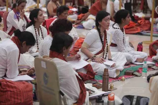 A seated group of women in white and cream clothing, many with red or orange shawls and rudraksha malas, sit on mats in a hall during a discourse session. Several write on loose paper with pens, one woman in the centre wears a white sari with gold border and a red shawl, a bindi on her forehead and a gold earring. Water bottles, small brass and wooden boxes, a brass lamp and a copper flask sit on the floor. A woman at left in a headset listens while another reads at the lower left. The scene reflects an attentive study session with notes being taken.