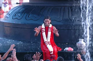 A photograph of a man in a red sleeveless garment and red shawl, wearing a white and red flower garland with red rose clusters, seated and smiling with arms raised as water pours over him from a large black polished stone lingam. Silver pot with flowers sits at right. Hands of devotees reach up from the bottom of the frame, and two figures in saffron robes stand at the upper left. a person: long dark hair, red attire and garland fit the presentation, though the face and build are not distinctive enough to confirm identity; treated as uncertain.