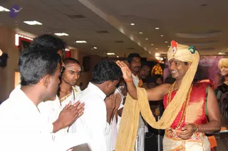 Indoor event photo in a bright hall with ceiling lights and a pink-and-white banner behind. On the right, a smiling man in a gold turban and a red and gold garment, wearing layered gold necklaces, a red bead mala and a gold waist sash, raises his right hand in blessing toward a line of men. Several men in white dhotis and shirts stand at left with palms pressed together in namaste; one has white tripundra marks on his forehead and a rudraksha-style mala. Other people stand in the background. a person in the turban and red garment, with gold ornaments and mala, giving blessings to devotees.