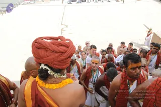 A photo of a group ascending stone steps toward a sandy riverbank. In the foreground, a figure seen from behind wears a bright orange-red turban decorated with white flowers, a marigold garland around the neck, and a saffron-orange shawl over bare shoulders. Around them, many devotees in saffron and red shawls, white dhotis and kurtas, and white-and-green lanyard badges climb the steps. Several men have orange tilak marks on their foreheads, and some wear rudraksha malas. A small figure in white and red stands far to the right on a raised platform. The bright sand and sunlight fill the background.