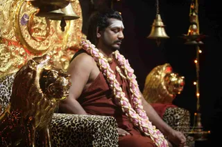 A seated man with closed eyes sits on a gilded throne flanked by golden lion heads and leopard-print cushions. He wears a rust-red robe, a double garland of pink and white flowers across his chest, and a white vertical mark on his forehead. Hanging brass bells and a lit oil lamp stand at right against a dark background.