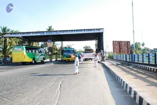 A roadside scene on a paved highway under a large metal gateway sign reading "EAST COAST CONSTRUCTIONS & INFRASTRUCTURE LTD" with "WELCOMES YOU" below. Two white-clad figures walk down the centre of the road, one carrying a white banner with red lettering. A green and yellow bus and a second yellow-green bus pass beneath the gateway, with palm trees and a blue-and-white railed bridge beside the road.