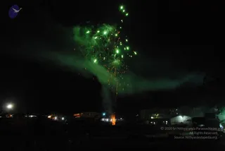 Night photograph of a green fireworks burst rising from a ground-level launch point, with bright sparks trailing down through dark sky and drifting smoke. Illuminated tents, a lit structure and a few distant lights are visible along a dark horizon at the base of the frame. No people are visible in the scene.