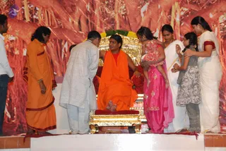 A photograph from a stage event: a man seated on a gilded throne with a red cushion, wearing an orange saffron robe with a red undershirt and bare feet resting on a golden footstool. Before him, a man in a light grey kurta bends forward, and a woman in a pink salwar kameez holds a small child in a red floral dress. A woman in white and a girl in a grey dress stand beside them, and on the left a man in a saffron dhoti and shawl stands near the stage edge. A red and orange backdrop with a floral garland on the throne. The seated figure's identity cannot be confirmed from appearance.