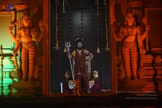 Night photograph of an illuminated temple-style entrance. Two large orange-lit guardian statues with multiple arms stand on either side. In the central niche, a man dressed in a maroon-red robe and wrap with a rudraksha mala and gold necklace holds a silver trident, standing beside a gold-framed throne. Strings of fairy lights and hanging bells frame the niche, and a small oil lamp burns at the base. The man is the only physical person visible; the statues are not people.