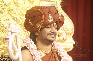 A smiling man seated before a gilded, ornately carved throne back, wearing a rust-brown turban decorated with a gold and jewelled brooch, a feather plume, and an orange beaded ornament on one side. He wears a pink and white flower garland, a gold chain necklace, a rust-brown robe, and a dark tilak mark on his forehead. A white ceremonial staff with a curved top stands at the left edge, and a microphone is at the right. A red curtain hangs behind.