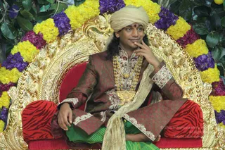 A man seated on an ornate gold throne decorated with yellow and purple marigold garlands and dark green leaves. He wears a beige turban with a red bindi, a maroon and gold embroidered long coat, a heavy gold necklace and crossed legs over a green cloth. His right hand rests at his chin in a contemplative pose, and the red and black tiger-striped throne cushions frame him. Swamiji (probable): long swept-back hair, a turban, ornate ceremonial gold necklaces and embroidered clothing, seated in a dais position.