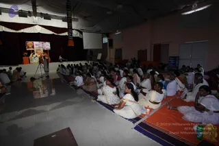 Wide photograph of a satsang hall with a large audience of women seated on orange and blue striped mats on the floor, many in white saris with coloured borders. On a raised stage at the back, a seated figure in white sits beside a man in orange robes standing near a microphone on a tripod. Behind them hang maroon drapes and white swags, and a banner reads Bhakti Spurana Program with a framed portrait. A projection screen and a blue notice board are visible at the side.