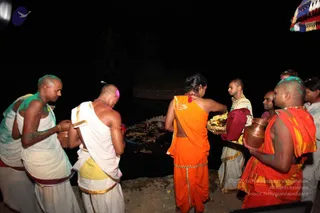 Night photograph at a water's edge with dark water behind a stone ghat. Several shaven-headed men in white dhotis and white upper cloths stand holding copper pots and brass vessels. In the centre a woman in an orange-saffron garment with gold trim carries a brass tray of flowers and offerings, and a man beside her in white holds a tray of marigold garlands. A man at right in orange-saffron dress with gold border and gold neck ornaments holds a copper pot, his head and face dusted with coloured powder. Floral offerings lie on the dark water surface. A red-and-white umbrella is visible at top right. Seven people are visible.