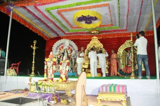 Outdoor evening ritual on a decorated stage under a multicoloured canopy with yellow, green, pink and orange borders and a central purple flower motif. A red backdrop frames an ornate golden throne-like seat garlanded with flowers, and a golden arch and a gold-framed deity image stand beside it. Several people in white dhotis and shirts gather around the central seat, one holding an object, while a man in an orange-and-saffron garment stands to the right. A deity image with many arms and a flame-like halo sits at left, flanked by two garlanded figures with orange and red garlands. Brass oil lamps (deepas) with flames stand on either side, and brass vessels and a kalasha-like pot sit on a gold-trimmed table in the foreground. A cameraman in a white shirt and jeans films the ceremony at right. Brass bells and a microphone are visible, and a small striped cushion sits on a gold stool.