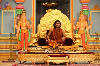 A seated man in orange-saffron robes sits on an ornate golden throne with lion-head armrests, draped with a tiger-striped cloth, centred in a temple-style hall. He wears a long rudraksha mala and a dark-brown beaded necklace, with long dark hair swept back, and smiles while resting his hands on his knees. Flanking the throne are two large painted deity murti figures with multiple arms, crowns and green-and-red dhoti-style lower garments, each holding ritual objects. Brass oil lamps stand on the golden floor, and a red carpet lies in front.
