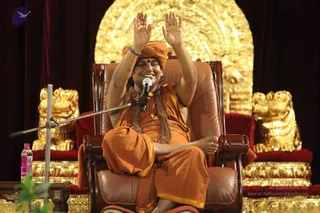 A seated figure in an orange saffron robe and orange turban sits in a brown leather armchair on a stage, arms raised high with palms open, smiling broadly. A microphone on a stand is in front of him, and a pink-capped water bottle sits at the left edge. Behind him are ornate gold-coloured carved thrones and a large gold arched backdrop. A rudraksha mala hangs around his neck and a red bindi marks his forehead.