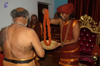 A photograph of an indoor room with red curtains and a gold-framed throne upholstered in deep red velvet with button tufting. A man in a red turban, red-and-gold dhoti and gold necklaces stands at right, smiling and holding up a long orange-red beaded garland over a woven basket. A bare-chested older man with grey hair, seen from behind, wears a gold-bordered cloth and a white sacred thread, and receives the garland. Two smiling men in red-and-gold robes and beaded necklaces stand in the background. The turbaned man wears distinctive gold ornamentation and a swept-back hairstyle consistent with the page's description, so he is labelled as a probable Swamiji.