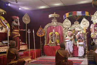 Photograph of a temple ritual inside a decorated hall with maroon walls and a purple spotlight. A standing man in a rust-orange dhoti and orange head cloth holds a tray of flowers and touches his forehead, while two men in white dhotis with garlands stand at a golden-framed shrine. A deity murti with a silver face and a garland of flowers sits under a fabric canopy with multicoloured tassels. Golden ceremonial umbrellas, a silver trident and tall decorated staffs line the stage. Seated devotees in rust-orange robes with flower garlands sit in the foreground, and a striped rug covers the floor scattered with petals.