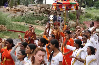 A festive outdoor procession moves along a dirt path past stacked flat stone slabs and green shrubs. A large ornate red, yellow and blue parasol with tasselled fringe is held above the group. A man in an orange robe with a rudraksha mala and a trident-topped staff stands among participants. Many young women and girls in saffron-orange and white robes with gold borders raise their hands and sing, some with white forehead markings. Several bald-headed men in orange and white clothing walk nearby, and a woman in an orange sari with a rudraksha mala stands at left. A man in white with a rudraksha mala is at right. Crowd members are visible in the foreground.