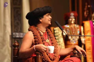 A man seated on a carved wooden throne-style chair holds a small white cup in his right hand, gazing to his left beside a microphone on a stand. He wears a large dark swept-back hairstyle, a dense multi-strand rudraksha mala over a red-orange shawl, a gold pendant, and green beaded necklaces. Behind him a gilded deity murti in ornate jewelled attire and a red crown stands on a dais, with orange and pink silk draped at right. Swamiji (probable) cues: swept-back hair, rudraksha mala, ceremonial gold necklace and saffron-red shawl; the page indicates he is present at this event.