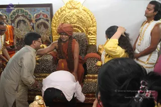A photograph of a seated figure on an ornate golden throne with a leopard-print cushion, backed by a golden arch and a painted panel of deities. The seated person wears an orange-red turban, orange robe, a long rudraksha mala and a shawl, and is being honoured. A man in a beige kurta leans forward and touches the seated figure's hand, while a bent devotee in white shirt and jeans touches the foot of the throne. A woman in a yellow top holds an apple-like fruit and touches the seated figure's head area, and a man in white with a gold border and gold necklace stands at right. Orange-clad attendants with garlands stand at left. The scene suggests a darshan blessing. a person: the seated figure's orange-red turban, orange robe and long rudraksha mala fit a person candidate, but the face is not clear enough to confirm.