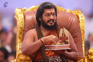 A bearded man with long swept-back dark hair sits in a gold-carved throne with brown leather upholstery, wearing a saffron-orange robe, a rudraksha mala with long beads, a gold necklace, a red bangle and a leopard-print cloth at the waist, with a red bindi on his forehead and a small headset microphone. He holds a small framed ceremonial figurine of a seated saint in orange robes and a gold-framed backdrop, set on a red and gold base, while speaking to a blurred crowd in the background. a person: the flowing hair, saffron robe, rudraksha mala, gold necklace and ornate throne match the page's a person reference, so this is an appearance-based candidate.