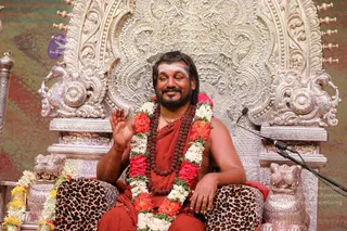 A seated man in rust-red robes sits on an ornate silver throne with carved peacock and lion motifs, wearing layered garlands of red and white flowers and a rudraksha mala. He raises his right hand in blessing, with a white vertical tilak on his forehead and a microphone stand beside him. The leopard-print cushioned seat and a wall of floral garlands frame the scene.