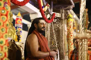 A bearded man with long dreadlocked hair stands in profile at a temple shrine, bare-chested under a loose saffron-orange wrap, with a vibhuti mark on his forehead and a red thread on his wrist. He holds a white cloth in his hands. Behind him are a silver-toned ornate pillar, red and orange flower garlands, and a tall garland column with marigold-like flowers. A second person in orange clothing with a rudraksha mala stands at the right edge. The scene suggests an evening puja at a temple setting.