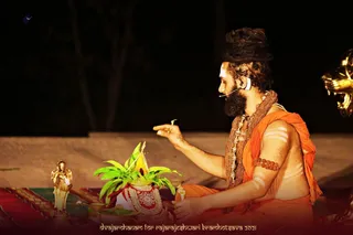A seated man in saffron-orange cloth performs a ritual at night beside a brass kalasha wrapped in red and white cloth topped with green mango leaves. He wears a large rudraksha mala and gold ceremonial necklaces, has white markings on his chest and arm, and holds a small lit item in his raised hand. A small gold deity figure stands on the left, a brass lamp sits near the pot, and a large gold ceremonial object is partly visible at the right edge. The caption along the bottom reads dvajarohanam for rajarajeshwari brahmotsava 2021. Visible appearance cues (long swept-back hair, saffron clothing, rudraksha and gold necklaces) match the page's note that Swamiji attended this event, so this is a probable identification.