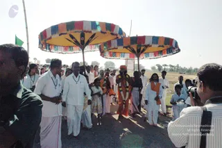 Outdoor procession on a dusty open field under a hazy sky with palm trees behind. Two large umbrella-style canopies with yellow, red, blue and green striped fringed panels are carried over the central figure. A man in orange robes with a tall orange head covering, a garland of flowers around his neck and a saffron shawl walks in the middle, appearing to be a person in a procession, with long dark hair and an orange shawl and garland as cues. Around him, devotees in white dhotis and white shirts, some wearing rudraksha-style neck malas, walk alongside. A child in a pale dress stands at the left of the group. A photographer in a striped shirt with a camera is at the right edge, and a green flag is visible at the left.