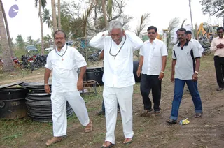 A photograph of a relief site outdoors among palm trees and scattered debris. In the centre, a man with swept-back grey hair, a grey moustache and a rudraksha mala stands in white shirt and white trousers, hands raised to his head. Beside him, a man in a white shirt and white trousers with a pendant necklace stands on the left. Other men in white or grey shirts and dark trousers stand nearby, and large black cooking pots and stacked metal plates lie on the ground. Motorbikes and parked vehicles are visible behind them.