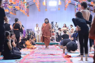 A photograph of a large hall with a white ceiling and hanging orange and multicoloured paper lanterns. A bearded man with long swept-back dark hair, wearing an orange-red shawl over an ochre robe and carrying a long staff, walks barefoot across a colourful striped mat toward a green-topped platform. The figure is an appearance-based a person candidate, identified by long hair, saffron-toned clothing and a staff rather than by face. Around him, participants in black clothing sit with palms pressed together in namaste, some kneeling or bowing with heads to the floor. A man in a white kurta with an orange-and-white garland stands with hands folded near the platform. A child in an orange outfit sits among the seated devotees. Water bottles and small cups sit on the mats, and a camera operator is visible in the background.
