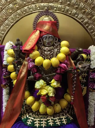 A dark metal deity murti of Sri Kalabhairava is displayed in a temple shrine with a golden arched backdrop. The murti wears a red and gold brocade headcloth, a gold and jewelled necklace, and a rudraksha mala, with a red and gold cloth draped over its shoulders. Its many arms hold a trident and other implements, and it is encircled by a garland of yellow lemons, pink roses and yellow flowers, framed by white jasmine garlands. No people are visible.