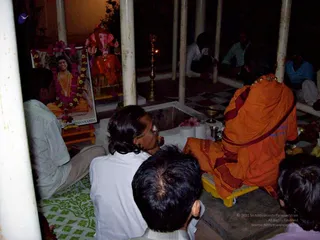A night-time ritual in an open-sided pavilion with white pillars and a tiled floor. In the upper left, a framed portrait labelled "Paramahamsa Nithyananda" is garlanded with pink flowers and beads. Beside it stands a Ganesha murti in orange and red clothing. A brass oil lamp burns in the centre, and a square fire pit with offerings sits in front. A person in saffron robes sits cross-legged on a yellow stool at right, facing the altar. In the foreground, several seated devotees in white shirts and dark-haired heads are seen from behind, and more people sit in the background among the pillars. The scene suggests a Ganesh Chaturthi puja.