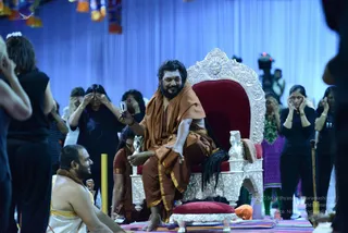 A bearded man in an orange-brown shawl and saffron-gold dhoti sits on an ornate silver throne with a red velvet seat and carved lion-head arms, raising a small handheld device in one hand while gesturing with the other. A seated priest in white with a red-brown cloth around his shoulders sits at the lower left beside a striped rug. Around the throne, several women in black tops stand with hands raised to their faces in a meditative pose, and a woman in purple and another in green stand behind, with a blue draped backdrop and colourful hanging decorations above. A cameraman with a camera is visible at the upper right. The bearded man wears a rudraksha-style mala, has long hair, and is seated in a throne in a manner consistent with the visible cues of a presiding teacher, so the identification is uncertain.